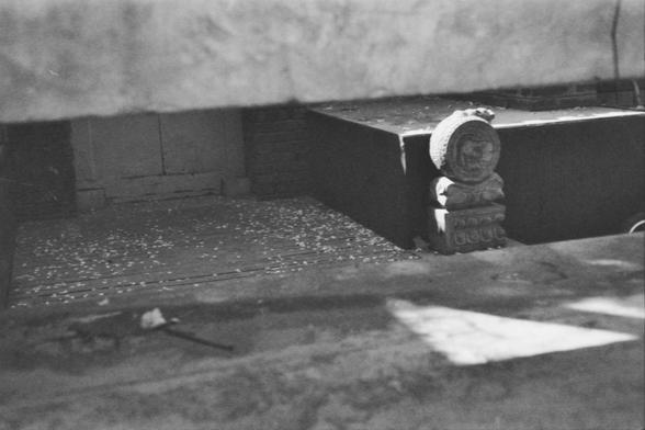 **English:**
A black and white photograph depicting a courtyard with a stone or concrete surface. In the center of the courtyard stands a stone pillar topped with a cylindrical stone. The ground is scattered with small debris. The courtyard is enclosed by high walls made of concrete or stone.

**Chinese:**
一张黑白照片，展示了一个铺有石头或混凝土地面的庭院。庭院中央有一个带圆柱形顶部的石柱。地面上散落着小碎片。庭院的四周是由混凝土或石头筑成的高墙。