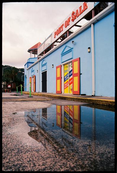 A film photograph of a long, blue, one-story building with a sign on its roof reading “port of sale” but the S is a dollar sign. The building has yellow doors and yellow and orange-red shutters. The building is reflected in a large puddle on the concrete street.