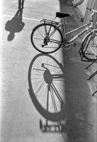 Monochrome photo. Sunny day. In the foreground is a bicycle that casts a shadow. A passerby's legs are seen in the background, from which a thick shadow is also falling on the asphalt.