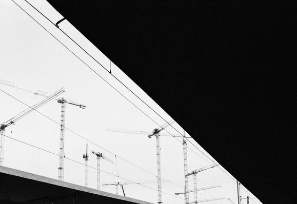 Black and white image featuring construction cranes in a skyline view. The top part of the image is dominated by a shadowy overhang, creating a stark contrast with the lighter sky. Wires are visible in the foreground, adding to the industrial atmosphere.