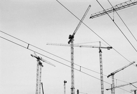 A black and white image featuring several construction cranes against a light gray sky. Power lines crisscross the scene, adding to the industrial atmosphere.