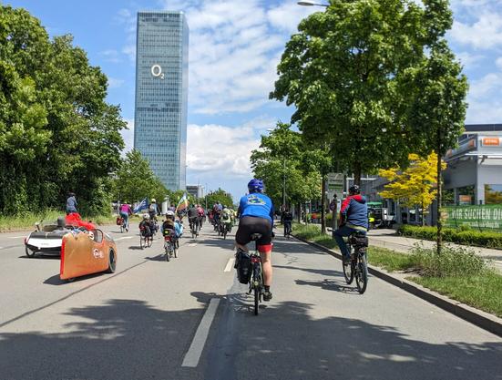 Vor der Kulisse des O2-Tower sind auf einer dreispurigen Straße keine Motorfahrzeuge unterwegs, nur Fahrräder in alen Varianten: links 3 Liegendräder mit Karosserie in Gelb, Weiß und Rot, dann eine Mischung von Liegend-, Stadt- oder Tourenrädern, z.T. mit Taschen, fast alle mit Helm. Rechts und links hohe grüne Bäume. Der Himmel ist blau mit einigen Wolken, der Vordergrund im Schatten.