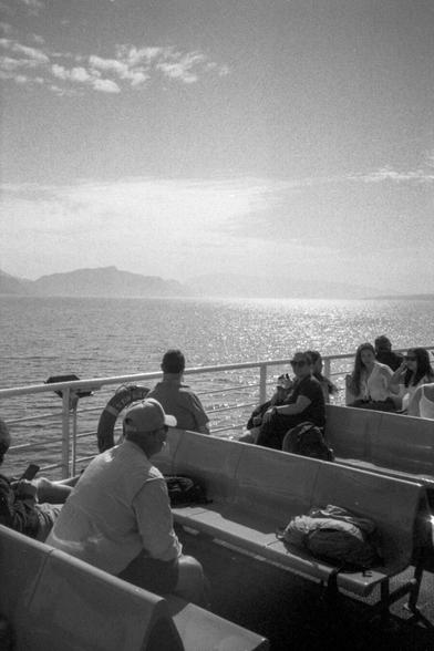 Passengers are riding on a ferry on the Croatian coast, enjoying a scenic view of the sea and distant mountains. The foreground shows rows of benches where people are seated, some facing forward and others turned to the side. A life preserver is attached to the ferry railing, adding to the maritime atmosphere.