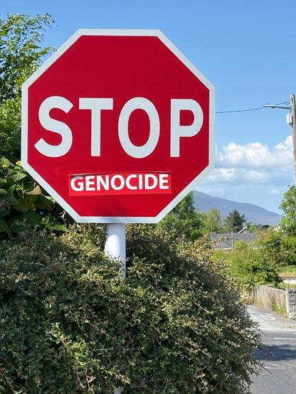 A red stop sign sticking out of bushes, a sticker reading "Genocide" in white letters has been added below the word "Stop" -  a road and a garden wall on the right and the edge of a mountain are seen in the background