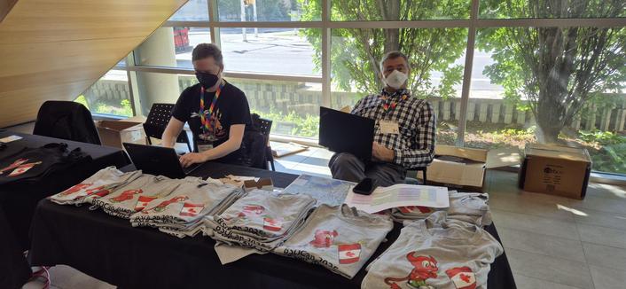 Two BSDCan organizers with one laptop each, sitting at the registration desk with the table covered in neat stacks of this year's BSDCan T-shirts