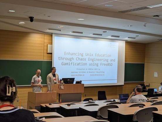 Photo taken during a talk at BSDCan 2025 titled “Enhancing Unix Education through Chaos Engineering and Gamification using FreeBSD” presented by Andreas Kirchner and Benedict Reuschling. The presenters are standing at the front of a lecture hall beside a large projection screen displaying the title slide. Groff is visible.