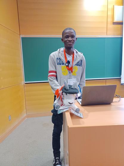 A smiling young Black man behind a podium, with a stuffed goat sitting on the podium