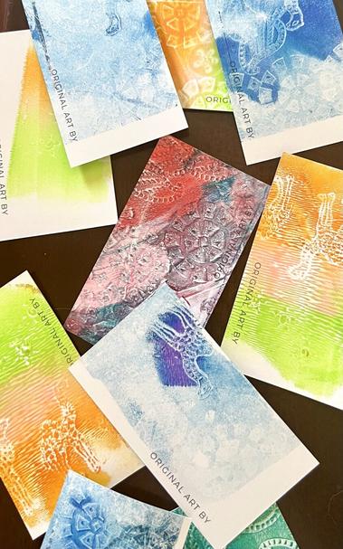 Business card-sized prints made using a gelli plate, acrylic paint and wood stamps. These are all different colours, some single colour, some many overlapping colours. Some motifs are visible; stamped, stylised leaves, flowers and a giraffe. The cards are scattered on a tabletop. These were pretty fun to make.