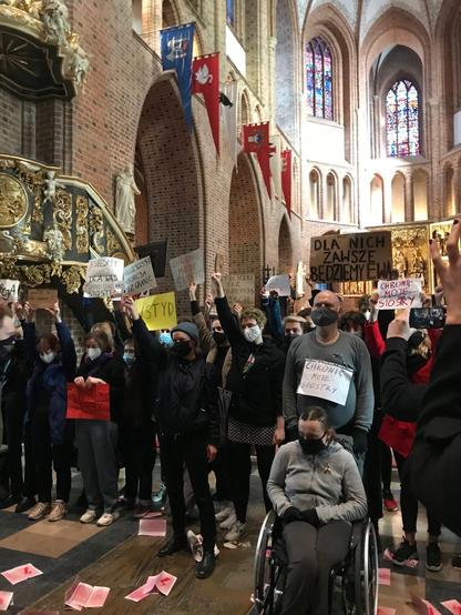 Wnętrze katedry R-K w Poznaniu, grupa osób w maskach z transparentami potępiającymi zaostrzenie zakazu aborcji. Na podłodze rozrzucone kilkanaście ulotek.