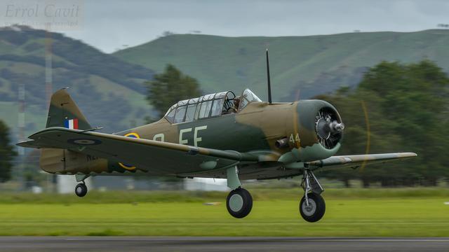A World War 2 trainer has just lifted off from a sealed runway, front canopy slid back. It has green and tan camouflage, matching the partially wooded hills beyond.