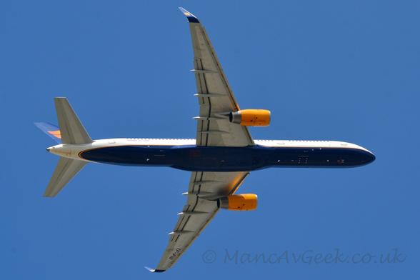 View from underneath of a twin engined jet airliner flying from left to right almost directly overhead, with flaps extended from the front of the wings, undercarriage already tucked away, and the nose raised sharply, suggesting it has just taken off.
The plane is mostly white, with a dark blue belly outlined with a yellow pin-stripe.
The engines mounted under the wings are a bright yellow.
The tail is also dark blue, with a pair of wavy yellow lines in the middle.
The up-turned wing-tips are also dark blue with a pair of bright yellow wavy lines.
The black registration "TF-FIO" can be seen under the outer sections of the left wing.
Vivid blue sky fills the rest of the frame.