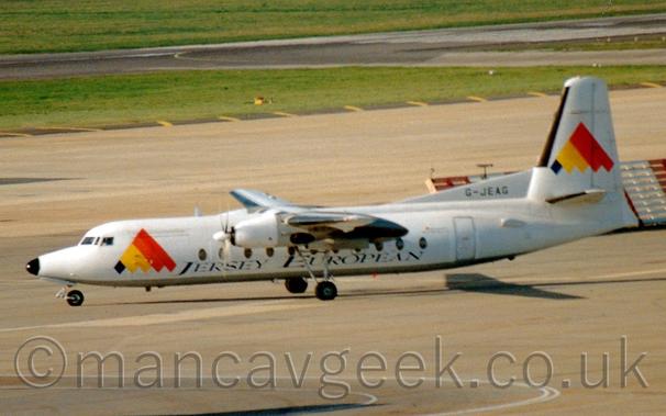 Side view of a high-winged, twin propellor-engined airliner taxiing from right to left, heading for the black runway visible behind them.
There are black "Jersey European" titles on the lower fuselage, with a large black, yellow, orange and red triangle on the forward fuselage just behind the cockpit, a logo repeated on the tail.
The black registration "G-JEAG" is on a white fillet at the front of the tail.
Grey concrete apron fills the foreground and a large part of the background, with large areas of green grass and a black runway filling the rest.