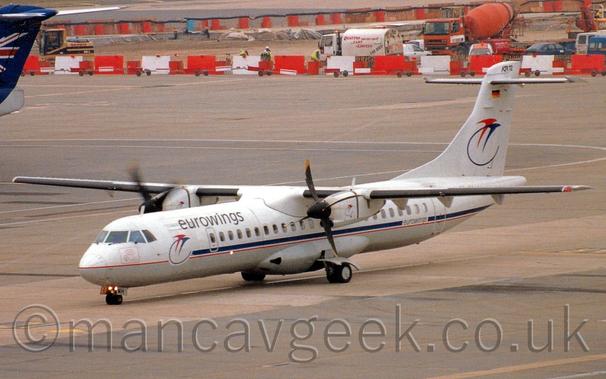 Side view of a high-winged, twin propellor-engined airliner taxiing from right to left.
The plane is mostly white, with a blue stripe and red pin-stripe running along the body, as well as a grey belly.
There are black "Eurowings" titles on the upper forward fuselage, above a pair of stylised flying bird, one red, one blue with a long tail that forms a semi-circle, a logo repeated on the tail.
Grey concrete apron fills the foreground and most of the background, with white and red barriers at the top of the frame denoting a construction zone, with several vehicles inside including an orange concrete truck, and men wearing hi-viz vests and hard-hats.