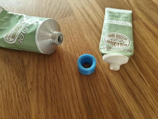 Two handcreme tubes, one travel sized, one refill sized with different screw top opening sizes and profiles as well as a 3d printed cylinder sitting on the table in-between of them.
The adapter fits the two screw tops and funnels from the larger to the smaller opening.