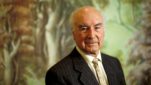 Elderly man in a striped suit with tie against a blurred forest-like background looking calm and composed.