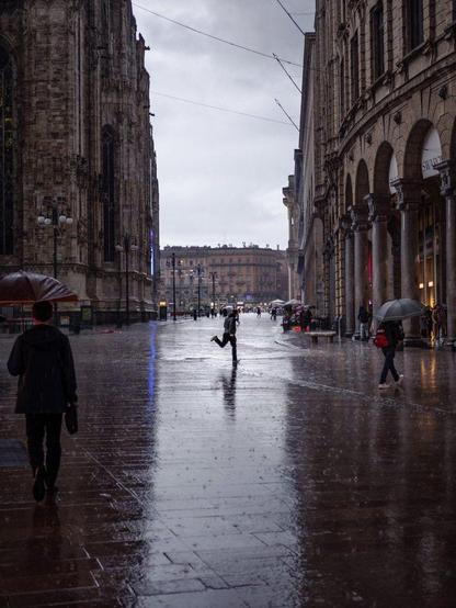 A man runs across a pedestrian street as it's raining. Other people are walking around the scene with umbrellas.