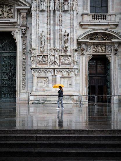 A yound woman walks on the Duomo's churchyard with a bright yellow umbrella. Her shape is reflected on the wet stone pavement.