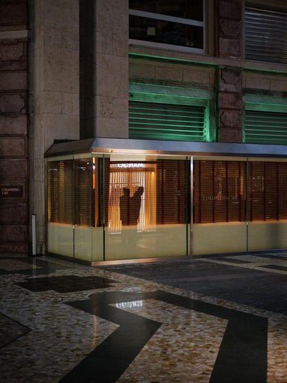 The silhouettes of two men are reflected into a restaurant terrace glass wall as yellow-orange light comes from a shop window outside the frame.
