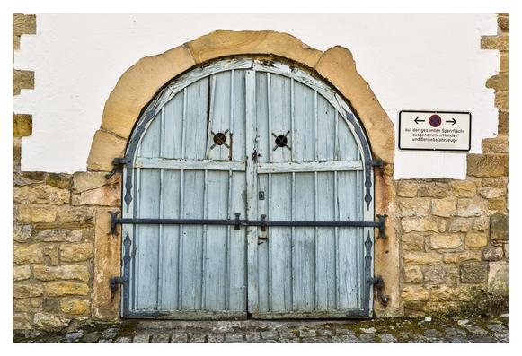 The picture shows an old, double-leaf wooden door in a pale shade of blue, set into a stone archway. The door has two small, round openings with metal grilles in the upper half and is reinforced with black metal hinges and locks. The archway is made of roughly hewn stones that are reddish-brown in color, while the surrounding wall is plastered white. To the right of the door is a sign with a traffic symbol indicating a no stopping zone in the vicinity of the door. The floor in front of the door is paved with uneven stones and there are some green moss patches around the edges. The door looks worn and gives the picture a historical charm.
