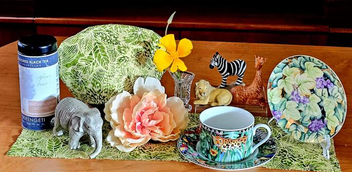 Tea tableau of an African scene with tropical fabrics and china, bright flowers, and a painted bisque collection of animals that includes an elephant, a zebra, a lion, and a giraffe. The tea is called Serengeti, a spiced black loose-leaf tea from Ethiopia.