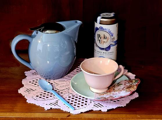 Simple and sweet tea tableau on a pink doily with a powder blue teapot, a pale green saucer, a blush pink cup, and sky blue teaspoon. There is a pastel floral fan-shaped napkin, and Earl Grey Macaron black loose-leaf tea from Pinky Up.