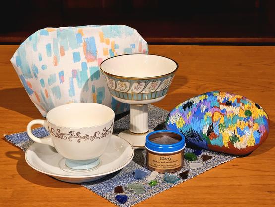 Tea tableau focused on splotches of color. The tea cozy is a fabric that looks like strokes of pastel paint; a large smooth rock is painted with a continuous cacophony of bright splashes of color; on the mat are scattered a handful of seaglass shards in a variety of colors. There is also vintage china and Cherry black loose-leaf tea.