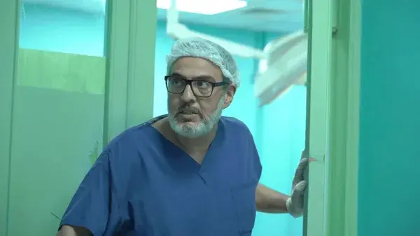 A still from the film A State Of Passion. A middle-aged man of Middle Eastern appearance, wearing medical scrubs and an urgent expression, steps out through a door into a hospital corridor.