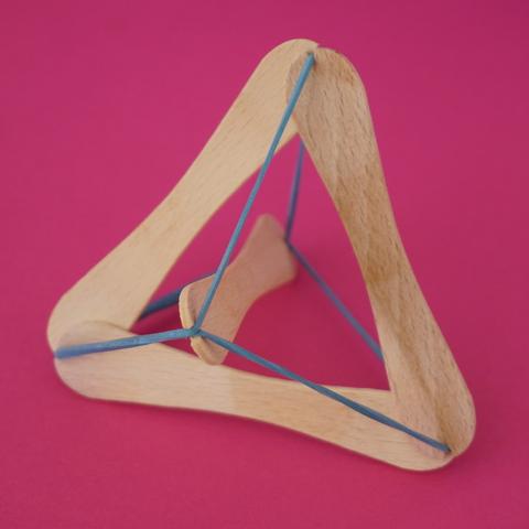 A tensegrity structure on a red background. It is made from four lolly sticks and two blue rubber bands. Three sticks form an equilateral triangle; the fourth stick is held by the rubber bands through the centre of, and perpendicular to, the triangle.