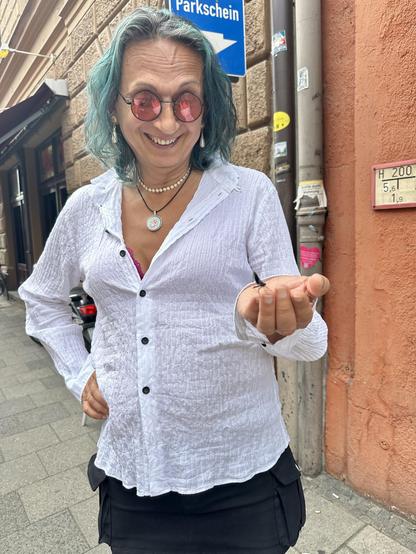 A person with teal hair, wearing pink sunglasses and a white button-down shirt, holds a small insect gently in their palm. They are smiling and wearing necklaces.  They are standing on a city sidewalk next to a building.