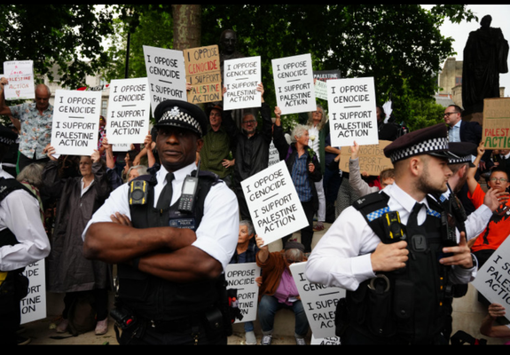 Two UK policemen stand in front of a protest in support of Palestine Action. The protesters have placard reading "I oppose genocide. I support Palestine Action"