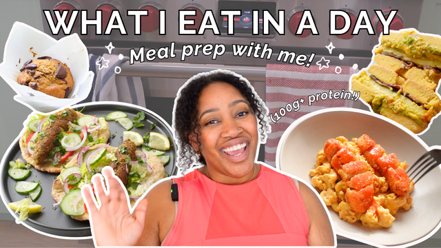 Thumbnail image for 'What I Eat in a Day' video featuring a smiling woman in a coral top waving, surrounded by photos of high-protein vegan meals including tacos, pasta with vegan shrimp, chocolate chip cookies, layered bars, and a modern kitchen stove in the background. Text reads: 'WHAT I EAT IN A DAY – Meal prep with me! – 100g+ protein!'