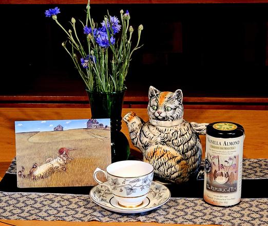 Tea tableau with a tabby cat theme. On an ornate black and beige cloth are set a striped cat figural teapot and an artwork of a tabby cat lazing on its back in a field of dry grass under a blue sky. There is vintage china, a vintage green glass vase holding deep blue cornflowers, and Vanilla Almond black tea from Republic of Tea.