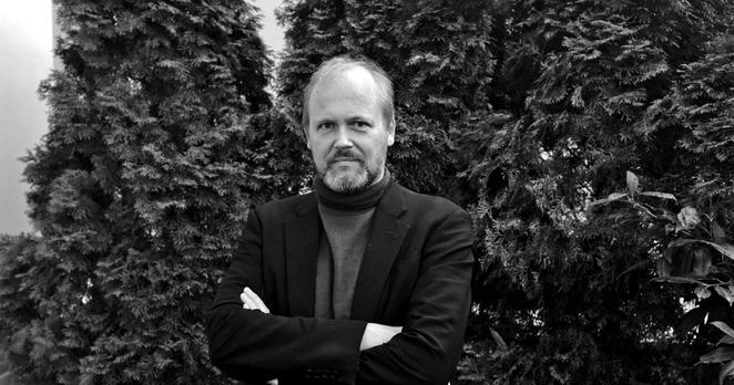 Schwarzweissfoto: Halbportrait eines Mannes um die fünfzig vor einer hohen Hecke.