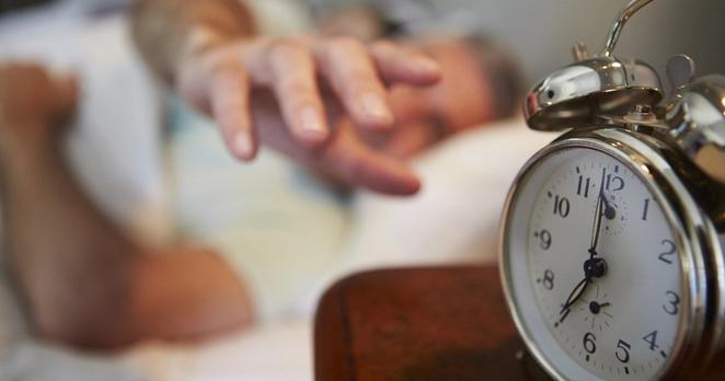 Im Vordergrund steht ein altmodischer Wecker und zeigt sieben Uhr. Unscharf sieht man dahinter einen Mann im Bett liegen, der die Hand ausstreckt, um den Wecker abzustellen.