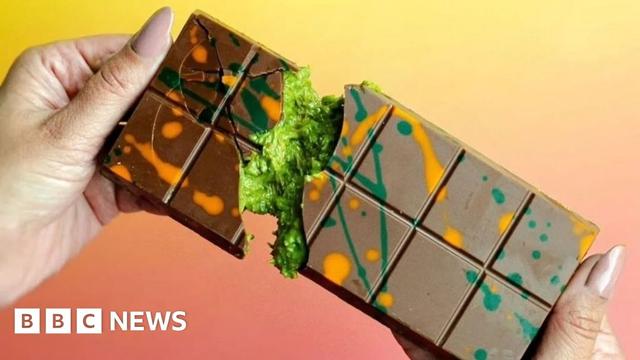 Hands snapping a bar of milk chocolate in half. The chocolate is covered in streaks of green and orange and, inside, a green, moss-like filling can be seen.
