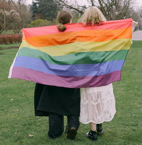Two women stand with their backs to the camera holding a rainbow LGBTQIA+ flag behind themselves.