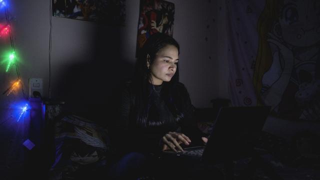 A woman sitting on a bed in a dimly lit room, focused on her laptop, with colorful fairy lights illuminating the surroundings, and various posters hanging on the walls.