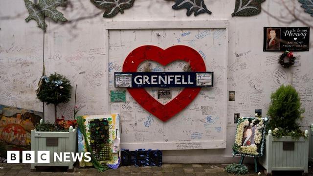 The memorial beneath Grenfell Tower, in west London. A red heart with the word "Grenfell" in white letters is mounted on a white wall covered in handwritten tributes to the dead.  There are small bushes in planters on either side of the image surrounded by other memorials.