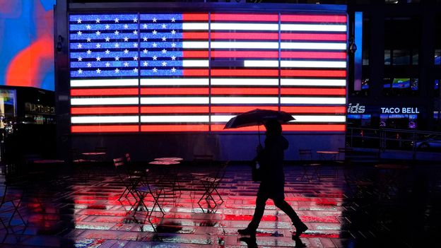 Person holding an umbrella walks in front of a Time Square screen showing a US flag (Credit: Getty Images)