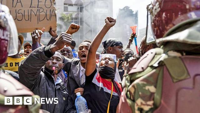 Protesters shout and whistle and raise their fists in front of police officers wearing riot gear near parliament in Kenya - 25 June 2024