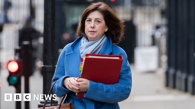 Lucy Powell seen wearing a blue jacket while holding a handbag and a red folder on the street.