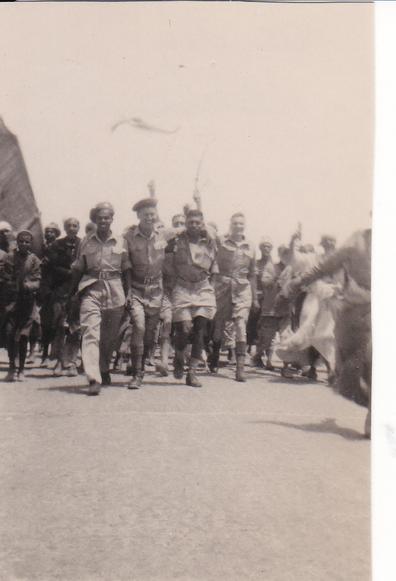Photo of three smiling men in service uniforms striding arm-in-arm towards the camera, closely followed by another waving a flag on a stick; and a small group of Egyptian civilian men, several of them also waving.