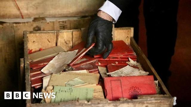 A person picks up a red booklet from a wooden crate. Other red booklets inside the box have a Swastika on the front. Green cards with handwritten notes can also be made out inside the box.