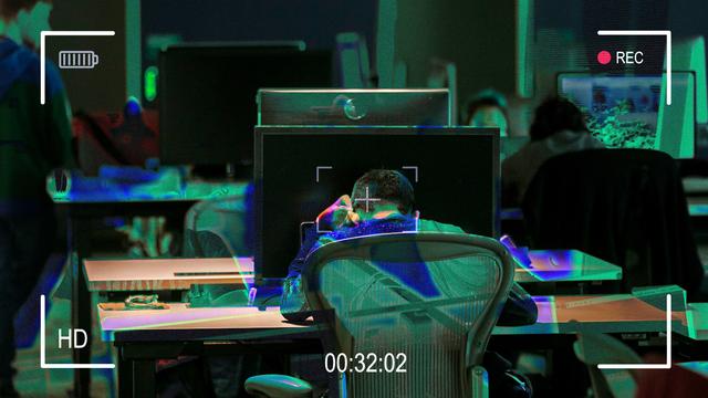 A person sitting at a desk in front of a large monitor, with their head resting on their hands, in a dimly-lit room filled with other workstations. Overlay features include a recording timer and battery indicator on the screen.