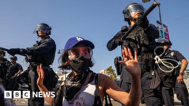A protester wearing goggles and a face mask holds their hands in the air, while officers stand over them