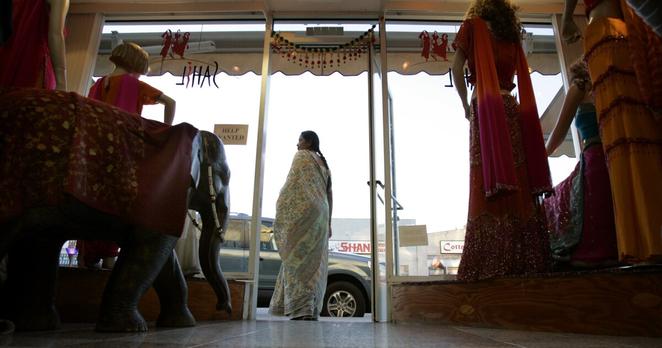 A woman in a floral sari stands in the entrance of an Indian clothing store, viewed from inside the shop. 