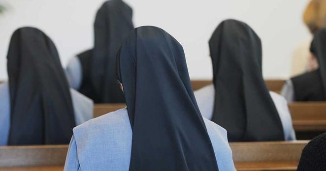 Das Bild zeigt eine Reihe von Nonnen, die in einer Kirche oder einem ähnlichen Andachtsraum sitzen. Sie sind von hinten zu sehen, und ihre Trachten sind gut erkennbar. Die Nonnen tragen einen traditionellen Habit, bestehend aus einem hellblauen Gewand und einem schwarzen Schleier, der ihr Haar bedeckt. Die Schleier sind lang und fallen in Falten über ihre Schultern. Ihre Haltung wirkt andächtig und respektvoll. Sie sitzen auf Holzbänken, die sich durch den Raum ziehen. Das Licht ist sanft und diffus, was eine ruhige und friedliche Atmosphäre erzeugt. Der Fokus liegt auf den Nonnen und ihrer gemeinsamen Hingabe. Die Komposition des Bildes lenkt die Aufmerksamkeit auf die Einheit und die gemeinsame Spiritualität der Gruppe.
