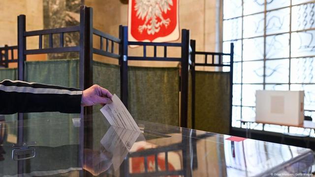 A hand drops a ballot into a ballot box. There is a paravent in the background. The Polish coat of arms is reflected in the surface of the ballot box