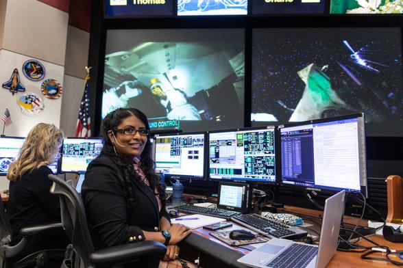 A woman sits at a desk and poses in front of three monitors. Multiple monitors display live data and video of astronauts on the International Space Station. An American flag and NASA mission patches are visible on the wall behind her.