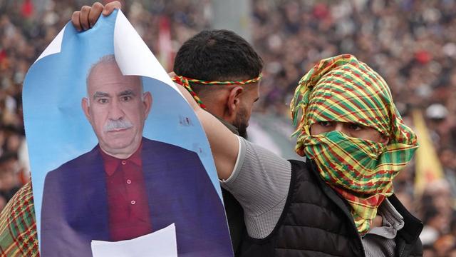 Zu sehen ist ein mit buntem Tuch vermummter Demonstrant. Dieser hält ein Poster hoch, das Abdullah Öcalan zeigt. 